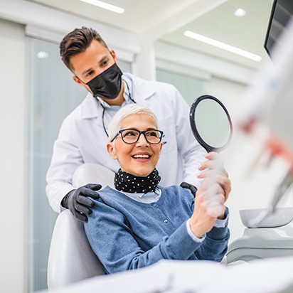 Woman in blue shirt in dental chair smiling at mirror with dentist behind her